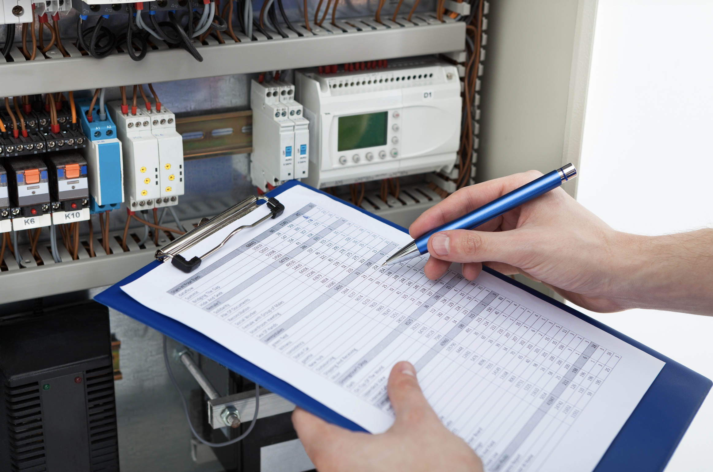 Close-up of an electrician inspecting an electrical control panel, holding a clipboard with a technical checklist and recording inspection results for an EICR